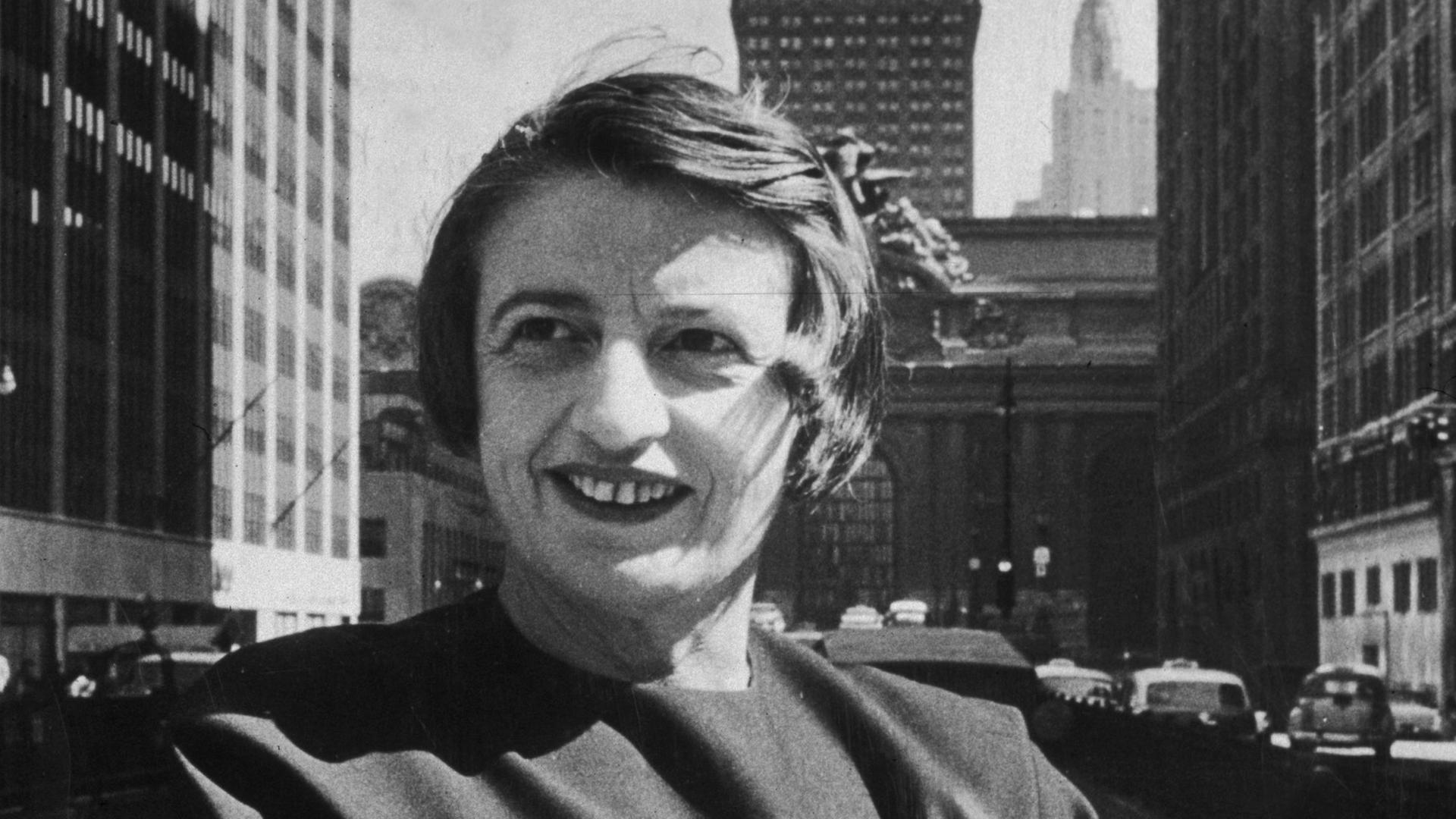 Porträt in Schwarzweiß von Ayn Rand, lächelnd und mit verschränkten Armen vor dem Grand Central Gebäude in Midtown Manhattan.