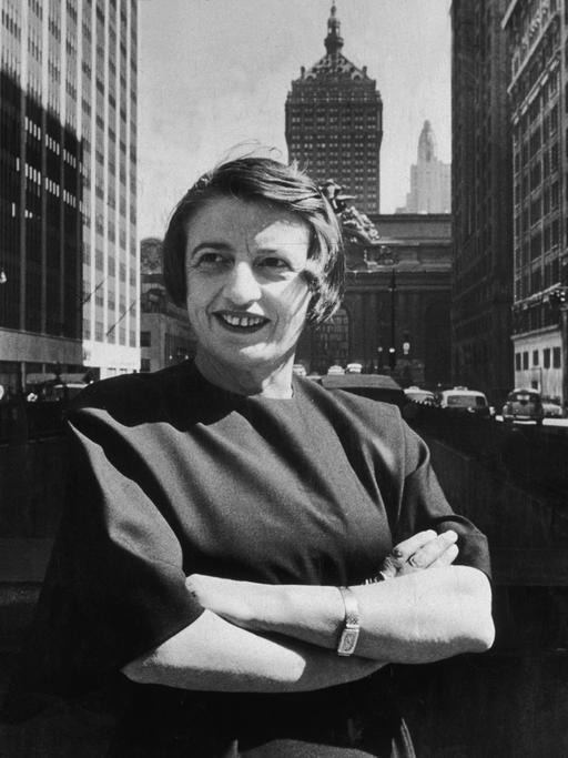 Porträt in Schwarzweiß von Ayn Rand, lächelnd und mit verschränkten Armen vor dem Grand Central Gebäude in Midtown Manhattan.