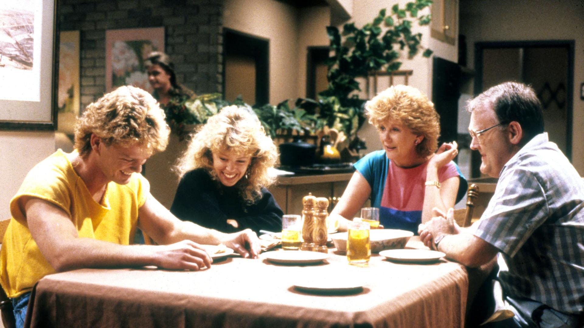 Szene aus der Serie "Neighbours", die Schauspieler Craig McLachlan, Kylie Minogue, Madge Charleston und Ian Smith sitzen gemeinsam am Küchentisch.