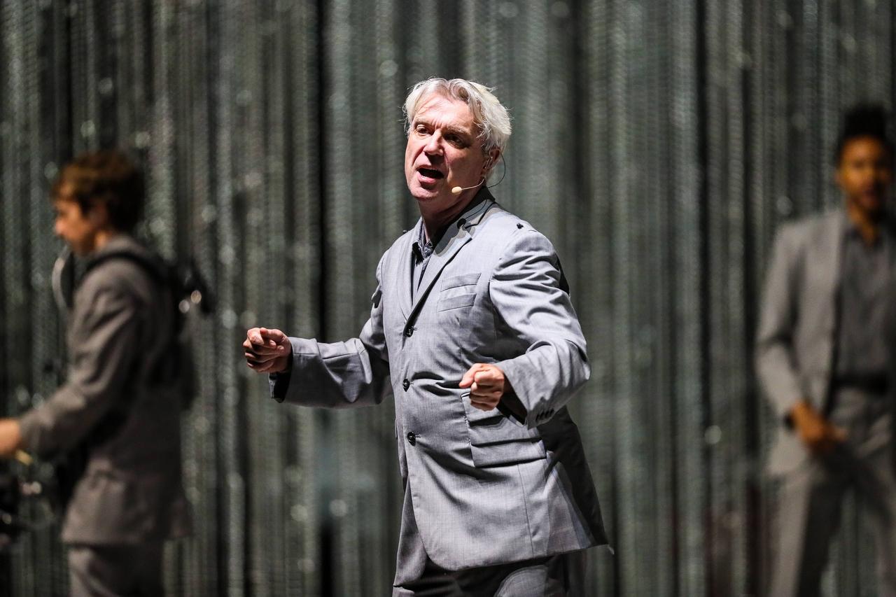 David Byrne steht auf der Bühne und bewegt sich. Er hat weiße Haare, trägt einen silbergrauen Anzug. Im Hintergrund ein dunkelgrauer Vorhang. David Byrne steht auf der Bühne und bewegt sich. Er hat weiße Haare, trägt einen silbergrauen Anzug. Im Hintergrund ein dunkelgrauer Vorhang.