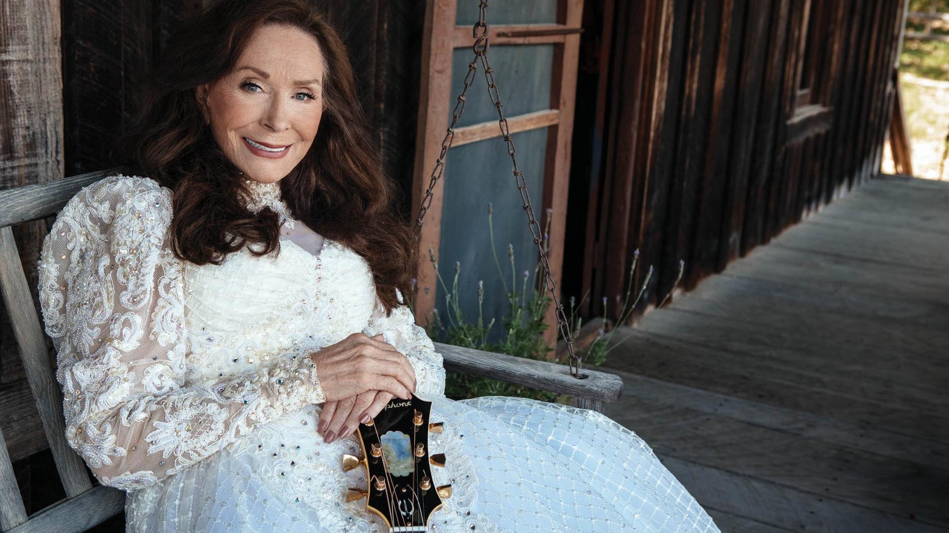 Countrylegende Loretta Lynn in einem weißen Kleid vor einem Holzhaus. Countrylegende Loretta Lynn in einem weißen Kleid vor einem Holzhaus.