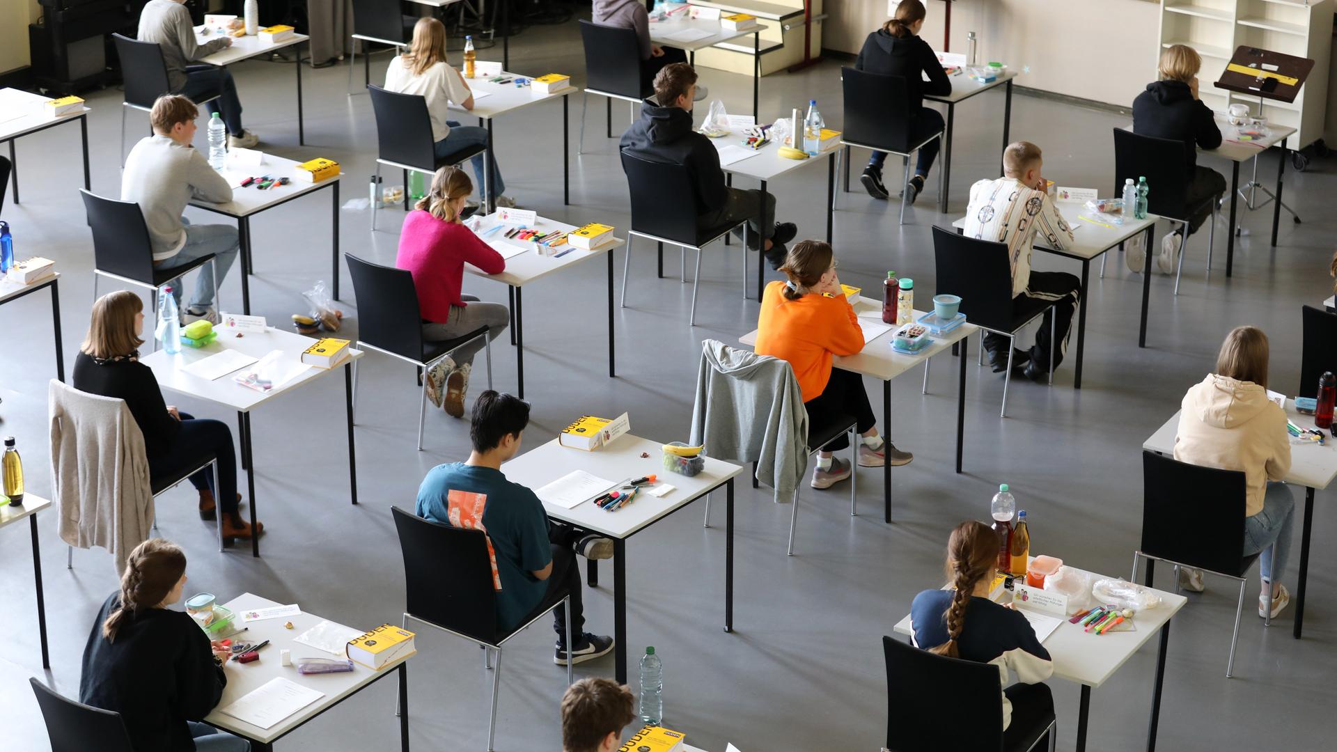 Jugendliche sitzen an Einzeltischen in einer Aula. Auf den Tischen liegen Schreibutensilien, Papier und ein Duden. Jugendliche sitzen an Einzeltischen in einer Aula. Auf den Tischen liegen Schreibutensilien, Papier und ein Duden.