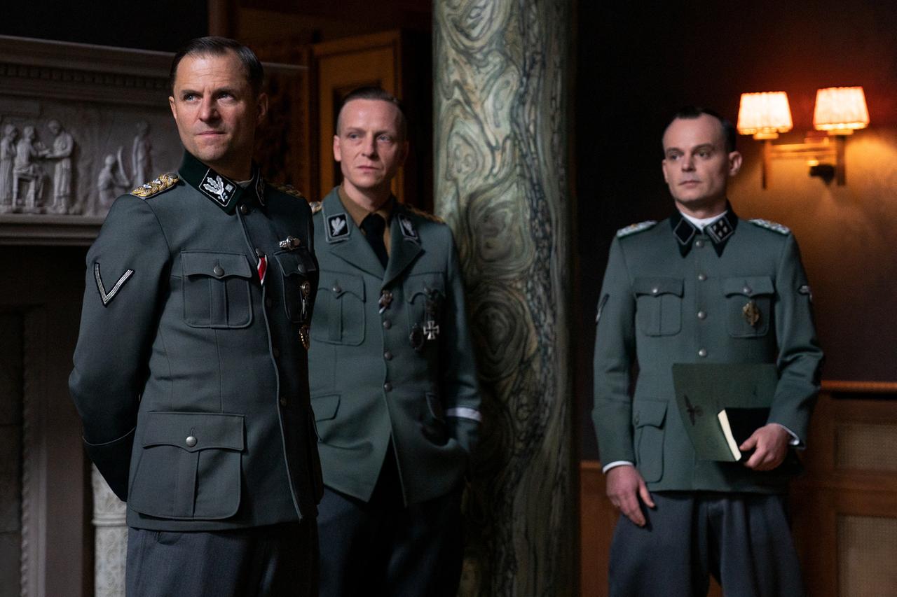 Filmstill aus dem ZDF-Film "Die Wannseekonferenz": Reinhard Heydrich (Philipp Hochmair), Heinrich Müller (Jakob Diehl) und Adolf Eichmann (Johannes Allmayer) stehen nebeneinander im Salon der Villa. Sie tragen Uniform, ihre Blicke sind ernst. Eichmann hält in seiner linken Hand Dokumente. Im Hintergrund des Raumes sind ein Marmorrelief und eine marmorierte Säule zu sehen. Filmstill aus dem ZDF-Film "Die Wannseekonferenz": Reinhard Heydrich (Philipp Hochmair), Heinrich Müller (Jakob Diehl) und Adolf Eichmann (Johannes Allmayer) stehen nebeneinander im Salon der Villa. Sie tragen Uniform, ihre Blicke sind ernst. Eichmann hält in seiner linken Hand Dokumente. Im Hintergrund des Raumes sind ein Marmorrelief und eine marmorierte Säule zu sehen.