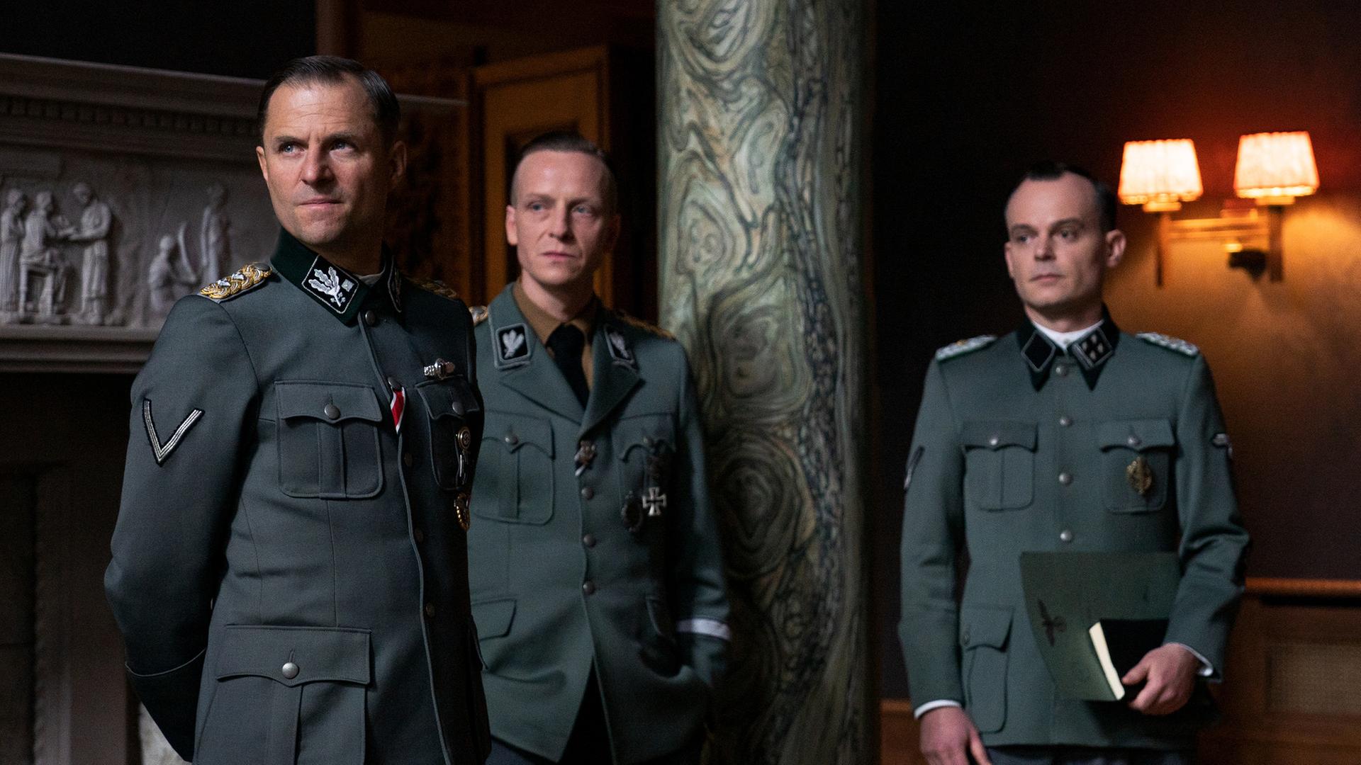 Filmstill aus dem ZDF-Film "Die Wannseekonferenz": Reinhard Heydrich (Philipp Hochmair), Heinrich Müller (Jakob Diehl) und Adolf Eichmann (Johannes Allmayer) stehen nebeneinander im Salon der Villa. Sie tragen Uniform, ihre Blicke sind ernst. Eichmann hält in seiner linken Hand Dokumente. Im Hintergrund des Raumes sind ein Marmorrelief und eine marmorierte Säule zu sehen. Filmstill aus dem ZDF-Film "Die Wannseekonferenz": Reinhard Heydrich (Philipp Hochmair), Heinrich Müller (Jakob Diehl) und Adolf Eichmann (Johannes Allmayer) stehen nebeneinander im Salon der Villa. Sie tragen Uniform, ihre Blicke sind ernst. Eichmann hält in seiner linken Hand Dokumente. Im Hintergrund des Raumes sind ein Marmorrelief und eine marmorierte Säule zu sehen.