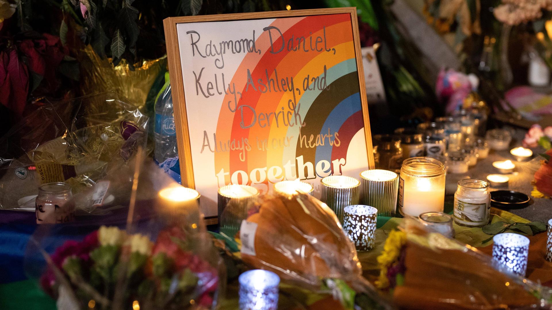 Ein gerahmtes Bild mit einem Regenbogen und den fünf Vornamen von Todesopfern steht an einem Trauerort mit Blumen und brennenden Kerzen in Colorado Springs.