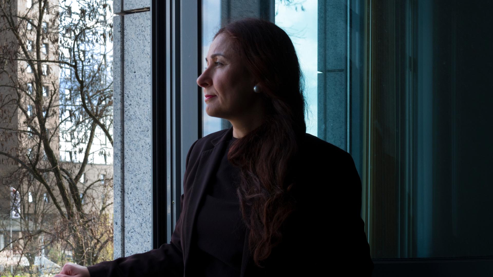 Sosan Azad, lange rotbraune Haare, Perlohrringe und lila Blazer, schaut aus dem geöffneten Fenster. Sosan Azad, lange rotbraune Haare, Perlohrringe und lila Blazer, schaut aus dem geöffneten Fenster.