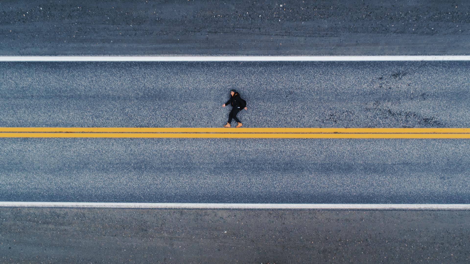 Luftaufnahme einer menschlichen Gestalt auf dem Asphalt liegend. Es wirkt auf den ersten Blick als würde die Person aufrecht laufen, statt flach auf dem Boden zu liegen. Unter den Füßen eine gelbe, horizontale Linie. Luftaufnahme einer menschlichen Gestalt auf dem Asphalt liegend. Es wirkt auf den ersten Blick als würde die Person aufrecht laufen, statt flach auf dem Boden zu liegen. Unter den Füßen eine gelbe, horizontale Linie.
