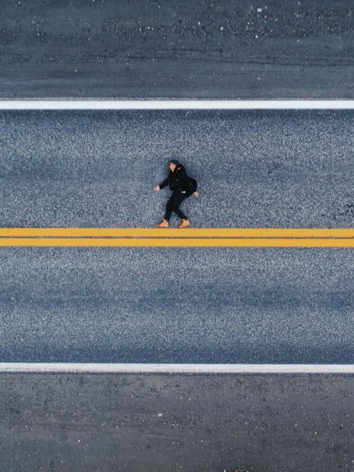 Luftaufnahme einer menschlichen Gestalt auf dem Asphalt liegend. Es wirkt auf den ersten Blick als würde die Person aufrecht laufen, statt flach auf dem Boden zu liegen. Unter den Füßen eine gelbe, horizontale Linie.