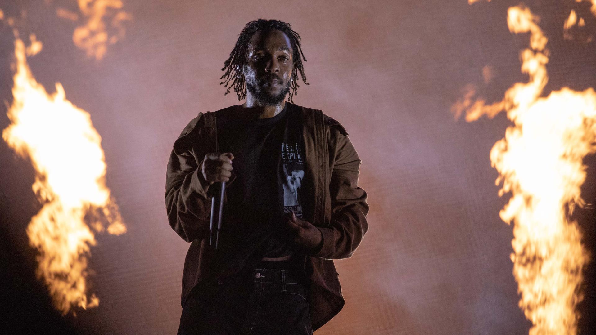 Der Rapper Kendrick Lamar steht auf einer Bühne, im Hintergrund sind Flammen zu sehen.