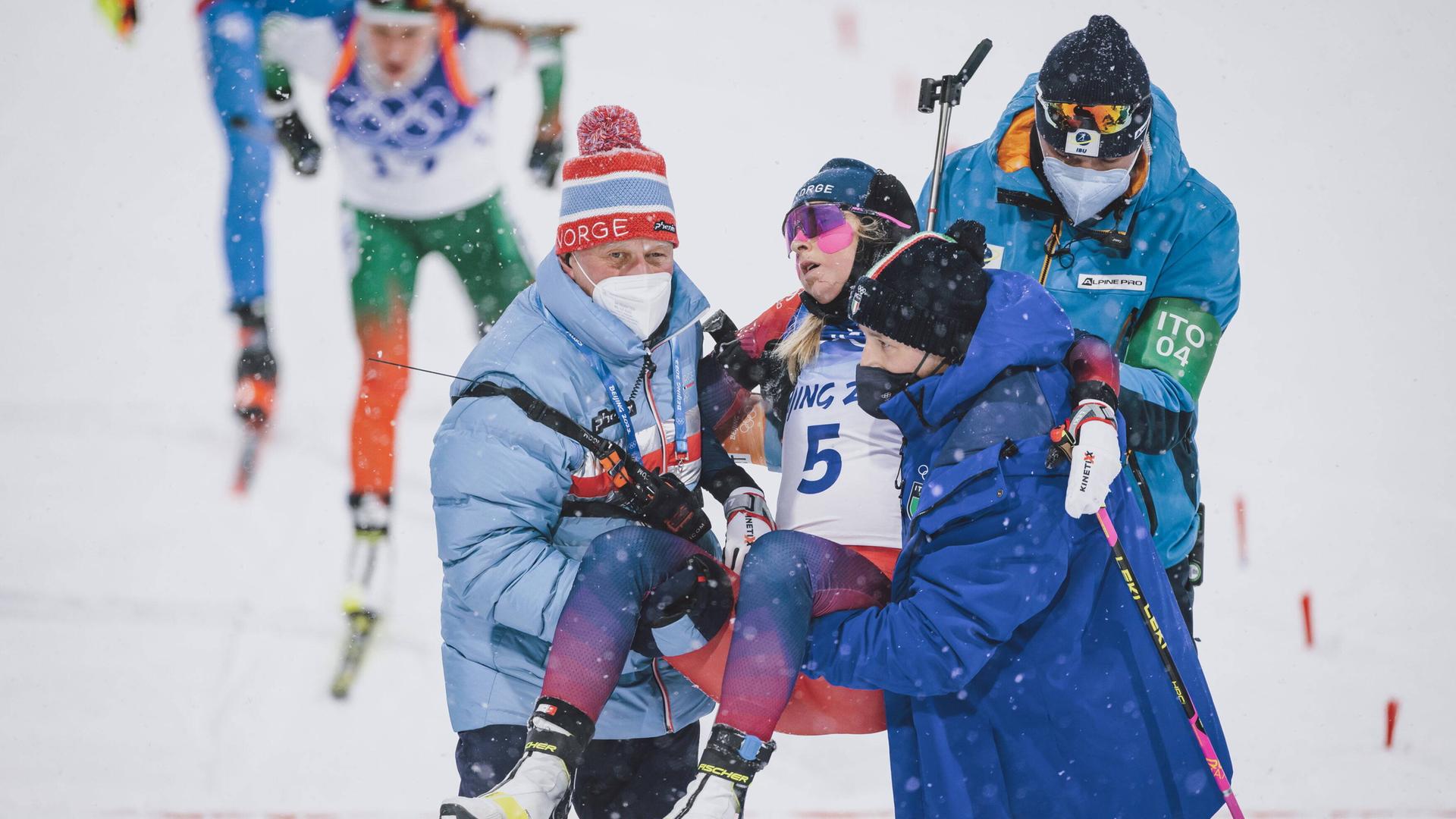 Die norwegische Biathletin Ingrid Landmark Tandrevold wird von zwei Helfern von der Piste getragen. Die norwegische Biathletin Ingrid Landmark Tandrevold wird von zwei Helfern von der Piste getragen.