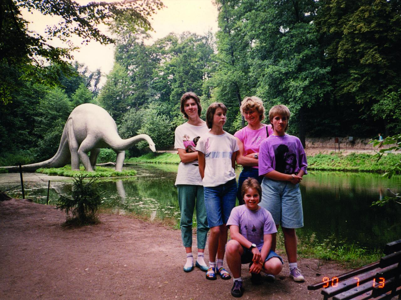 Teenager posieren vor einem Fluss. Im Hintergrund steht auf einer Insel das lebensgroße Modell eines Dinosauriers.