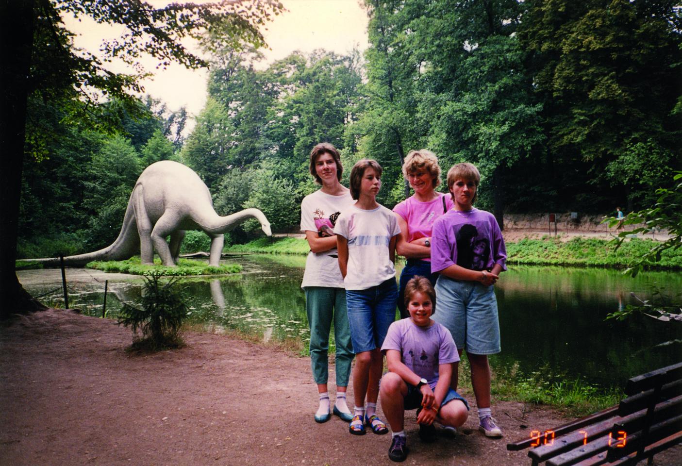 Teenager posieren vor einem Fluss. Im Hintergrund steht auf einer Insel das lebensgroße Modell eines Dinosauriers.