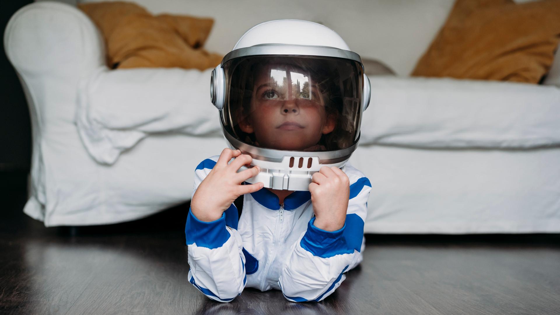 Ein Kind liegt in einem Astronautenkostüm auf dem Boden vor einem Sofa und guckt verträumt. Ein Kind liegt in einem Astronautenkostüm auf dem Boden vor einem Sofa und guckt verträumt.