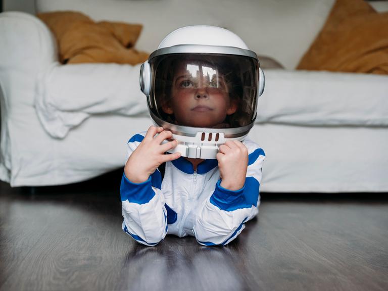 Ein Kind liegt in einem Astronautenkostüm auf dem Boden vor einem Sofa und guckt verträumt.