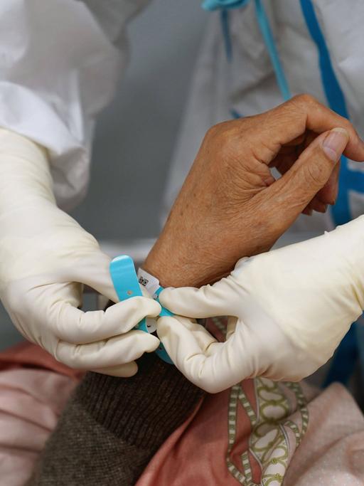 Ein Mensch in weißem Schutzanzug legt an die hand einer Patientin ein Armband an.