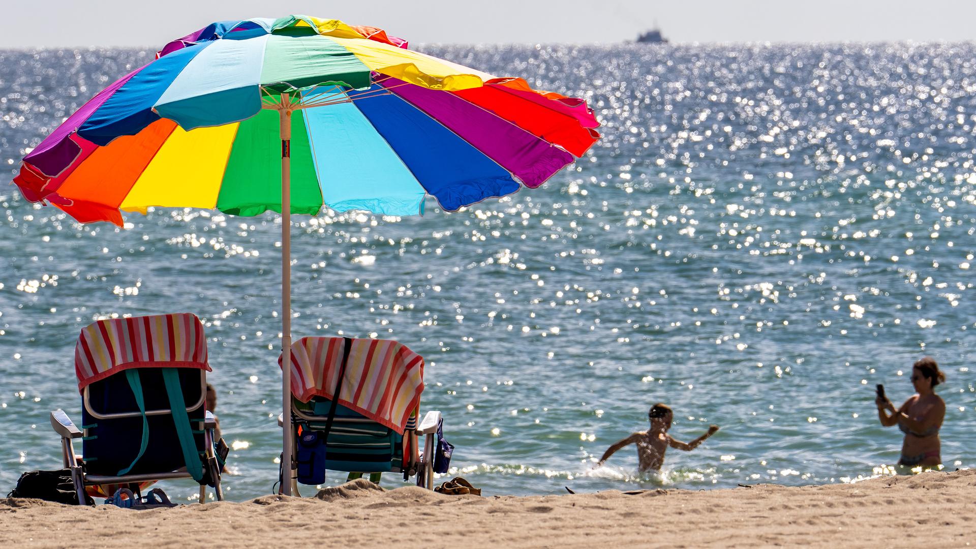 Ein bunter Sonnenschirm am Strand, im Meer planschen ein Kind und eine Frau Ein bunter Sonnenschirm am Strand, im Meer planschen ein Kind und eine Frau