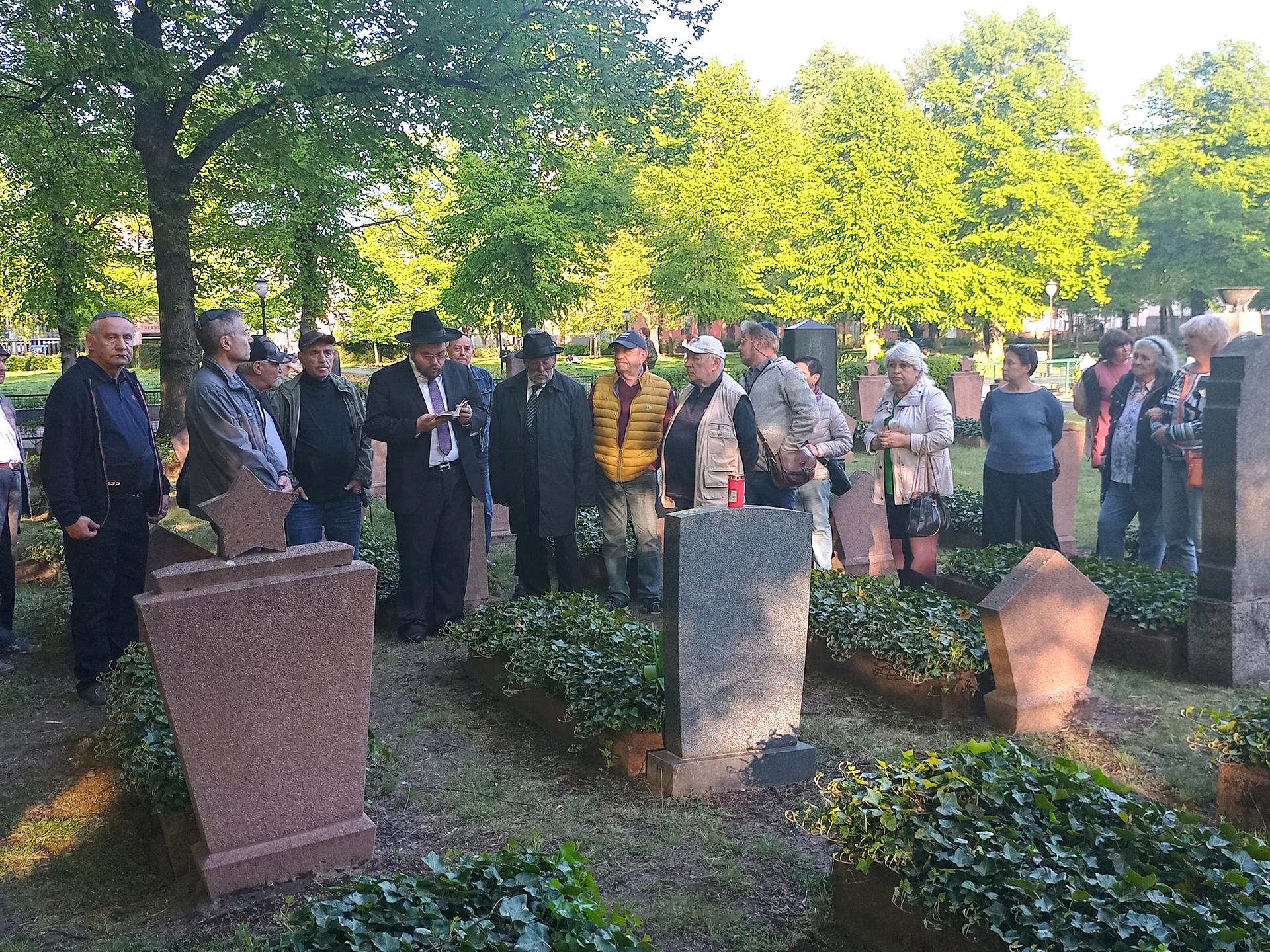Eine Menschengruppe und ein Rabbiner stehen auf einem Friedhof vor Grabsteinen. 