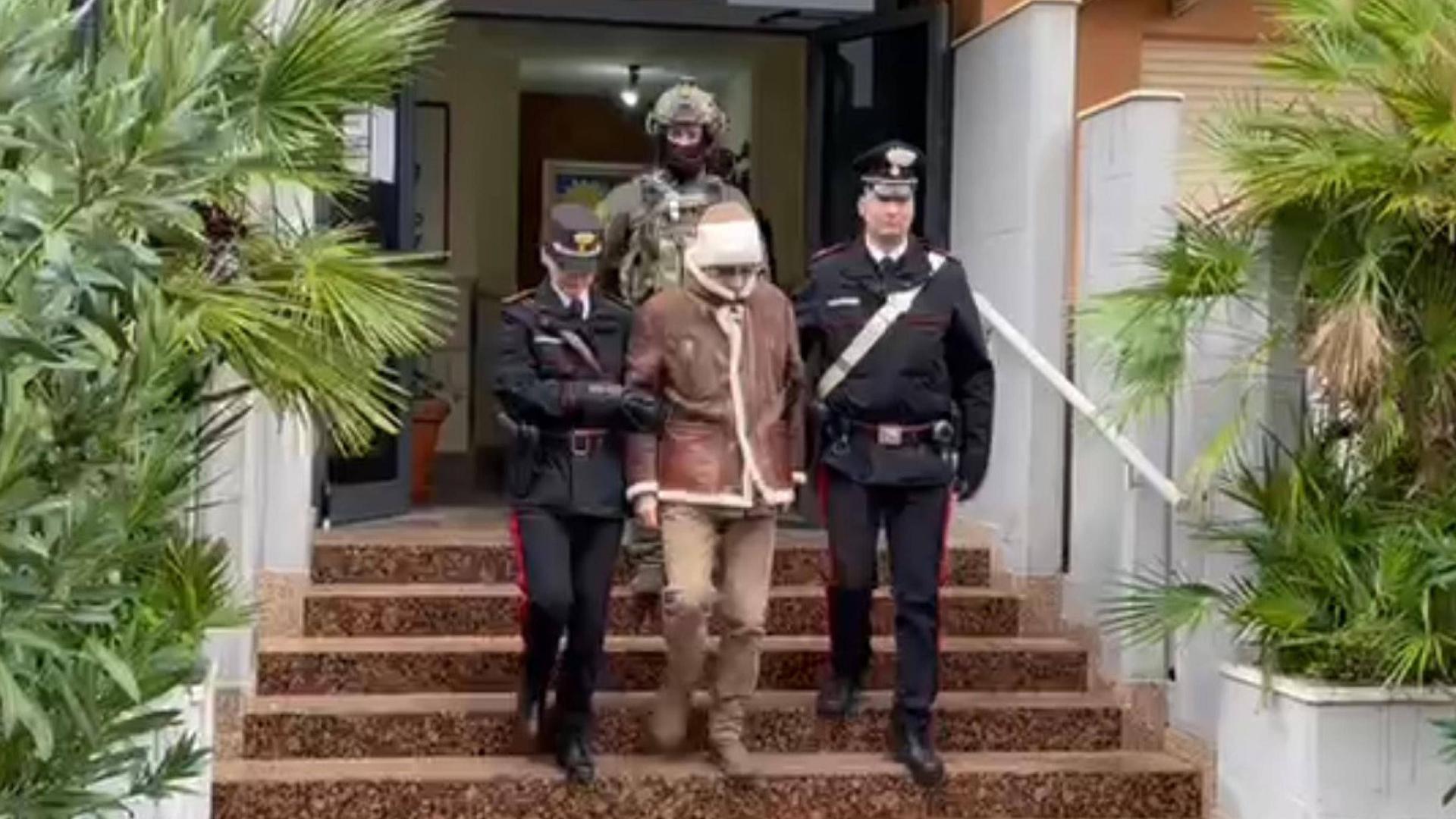 Der Mafiaboss Matteo Messina Denaro wird von zwei Polizisten eine Treppe hinabgeführt, hinter ihnen ein schwer bewaffneter Soldat. Der Mafiaboss Matteo Messina Denaro wird von zwei Polizisten eine Treppe hinabgeführt, hinter ihnen ein schwer bewaffneter Soldat.