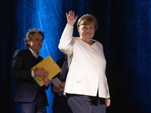 Angela Merkel. Eine Frau geht auf eine Bühne und winkt.