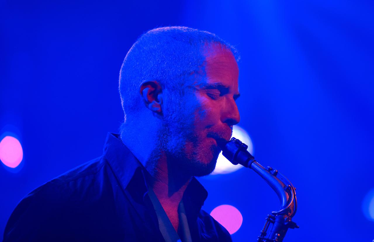 Ein Saxophonist mit extrem kurzen Haaren spielt sein Instrument, während er von blau-lila-farbenem Bühnenlicht umgeben ist. Ein Saxophonist mit extrem kurzen Haaren spielt sein Instrument, während er von blau-lila-farbenem Bühnenlicht umgeben ist.