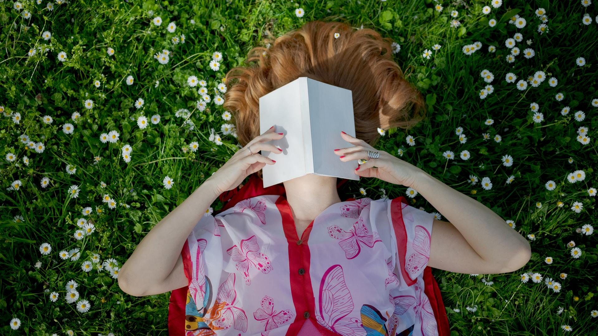 Eine Frau liegt im bunten Kleid auf einer Wiese voller Gänseblümchen und hält ein aufgeschlagenes Buch über ihr Gesicht.