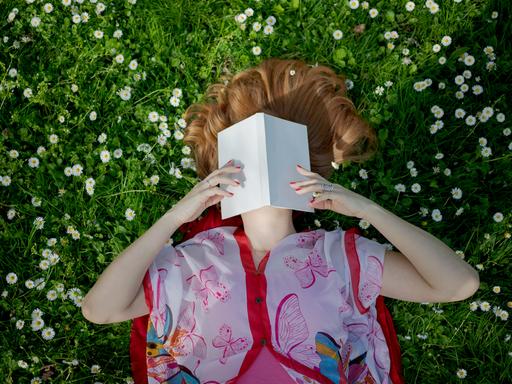 Eine Frau liegt im bunten Kleid auf einer Wiese voller Gänseblümchen und hält ein aufgeschlagenes Buch über ihr Gesicht.