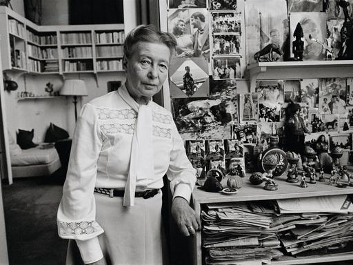 Simone de Beauvoir steht in ihrer Wohnung voller Bücher und schaut in die Kamera