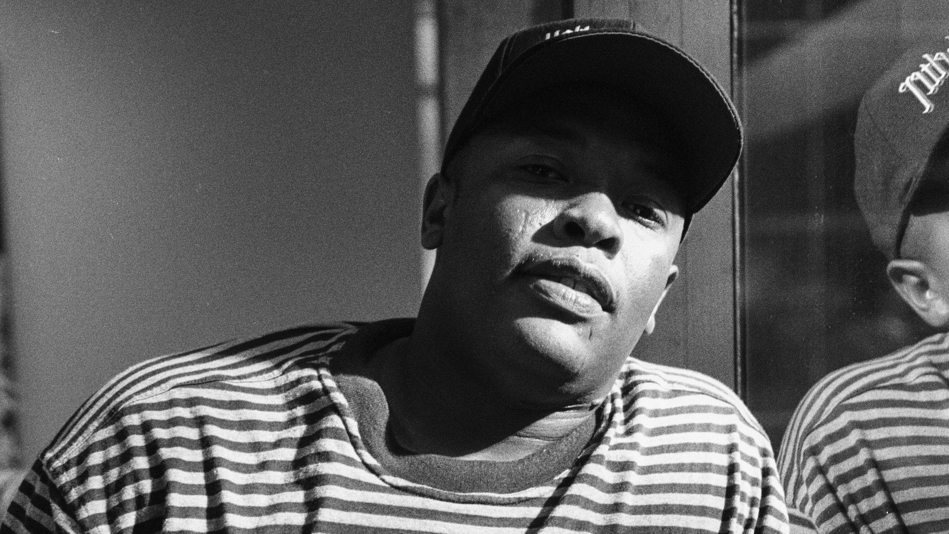 Schwarzweißfoto von Dr. Dre aus den 1990er Jahren. Er steht im Ringelshirt und mit Basecap an die Glaswand eines Tonstudios gelehnt und schaut direkt in die Kamera.