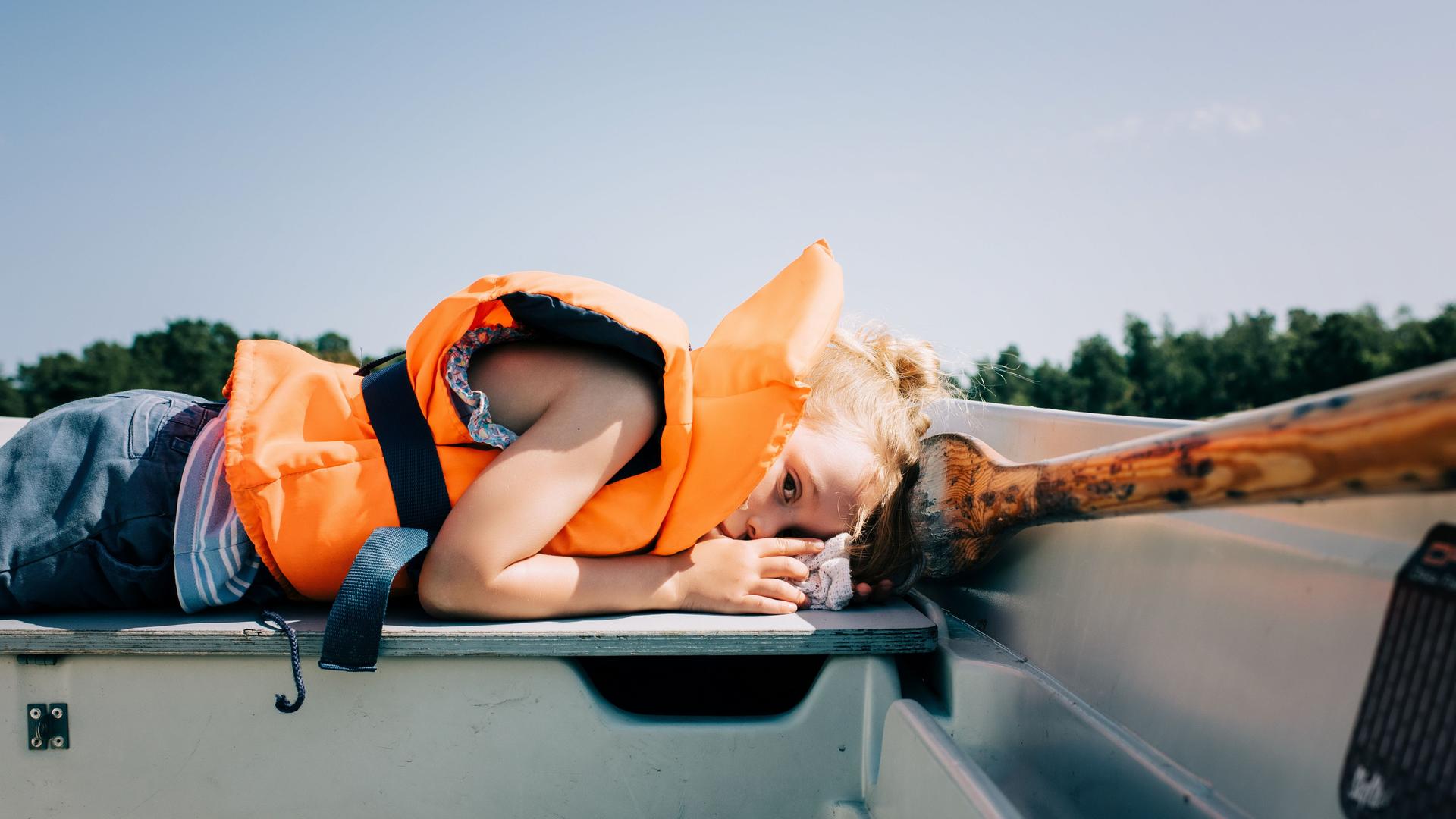 Ein Kind liegt in einem Ruderboot. Es trägt eine neonorangefarbene Rettungsweste. Ein Kind liegt in einem Ruderboot. Es trägt eine neonorangefarbene Rettungsweste.