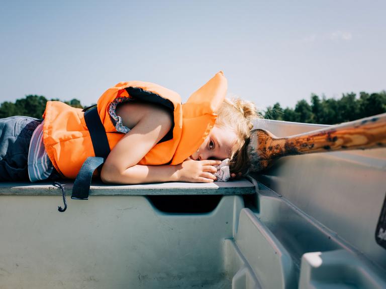 Ein Kind liegt in einem Ruderboot. Es trägt eine neonorangefarbene Rettungsweste.