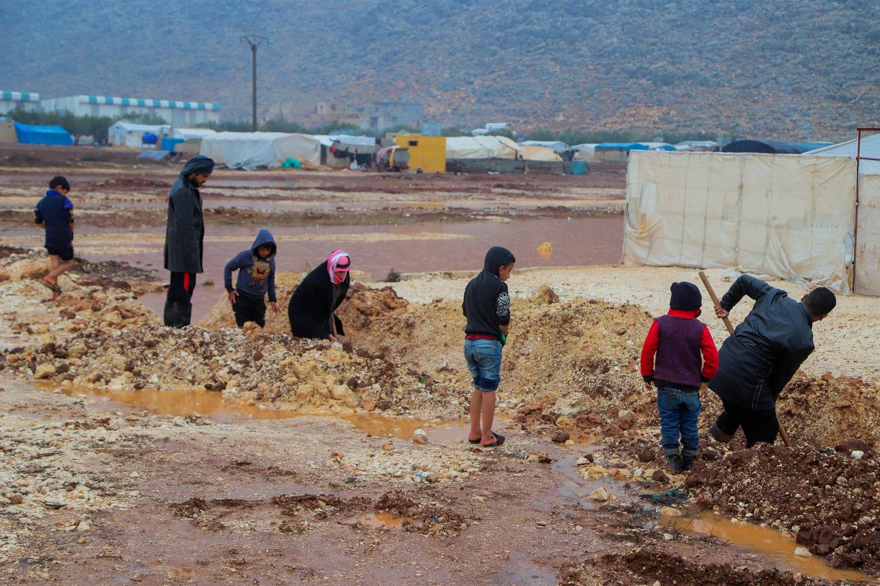 Kinder und Erwachsene graben in einem Flüchtlingscamp im Nordwesten Syriens im Matsch. Ein Kind steht in kurzen Jeans und Flipflops am Rand. Das Foto wurde im Dezember 2021 aufgenommen.