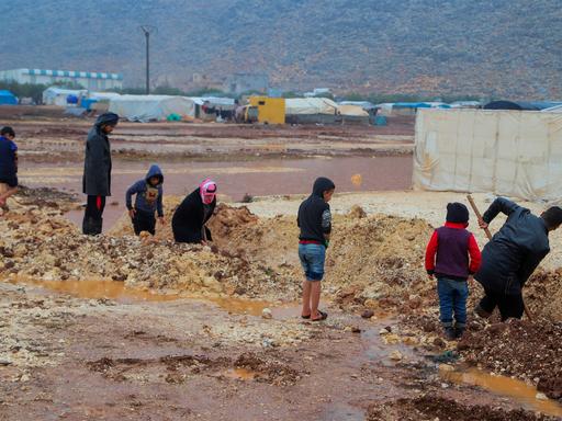 Kinder und Erwachsene graben in einem Flüchtlingscamp im Nordwesten Syriens im Matsch. Ein Kind steht in kurzen Jeans und Flipflops am Rand. Das Foto wurde im Dezember 2021 aufgenommen.