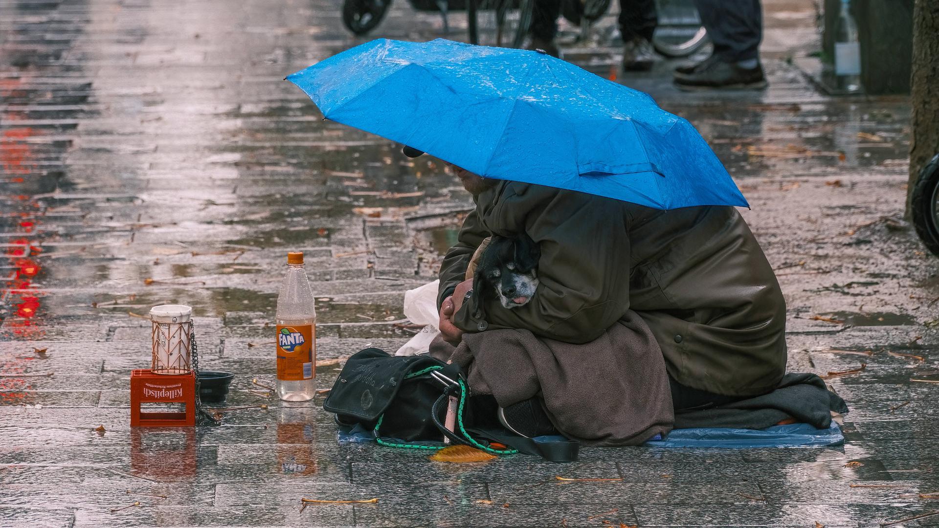 Ein Obdachloser sitzt mit seinem kleinen Hund auf einem verregneten Gehweg. Ein Regenschirm bietet den beiden notdürftigen Schutz. Ein Obdachloser sitzt mit seinem kleinen Hund auf einem verregneten Gehweg. Ein Regenschirm bietet den beiden notdürftigen Schutz.
