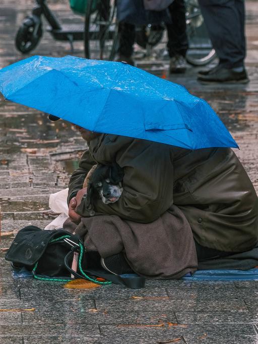 Ein Obdachloser sitzt mit seinem kleinen Hund auf einem verregneten Gehweg. Ein Regenschirm bietet den beiden notdürftigen Schutz.