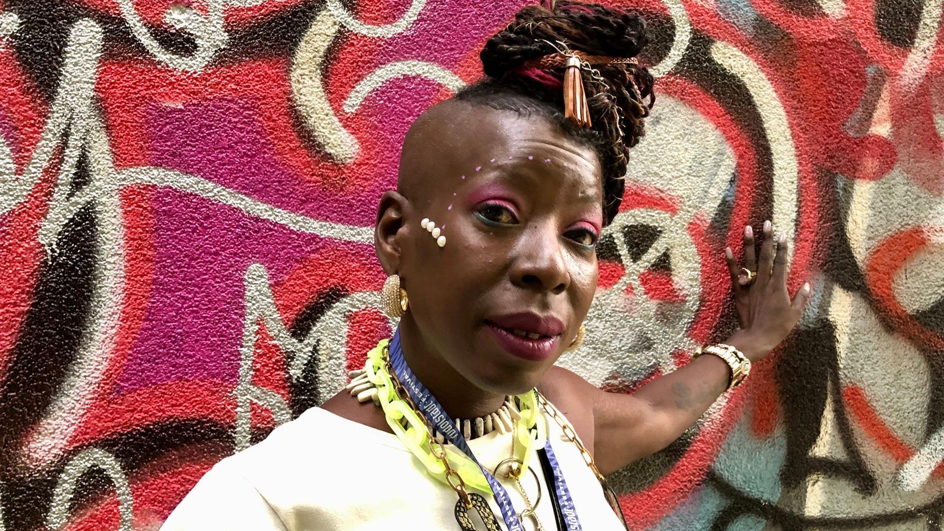 Eine Frau steht vor einer bunt besprayten Wand. Sie lehnt mit ihrer linken Hand gegen die Wand. Ihre Haare hat sie kunstvoll in die Höhe frisiert. Sie trägt auffälliges Make-Up und Schmuck. Eine Frau steht vor einer bunt besprayten Wand. Sie lehnt mit ihrer linken Hand gegen die Wand. Ihre Haare hat sie kunstvoll in die Höhe frisiert. Sie trägt auffälliges Make-Up und Schmuck.