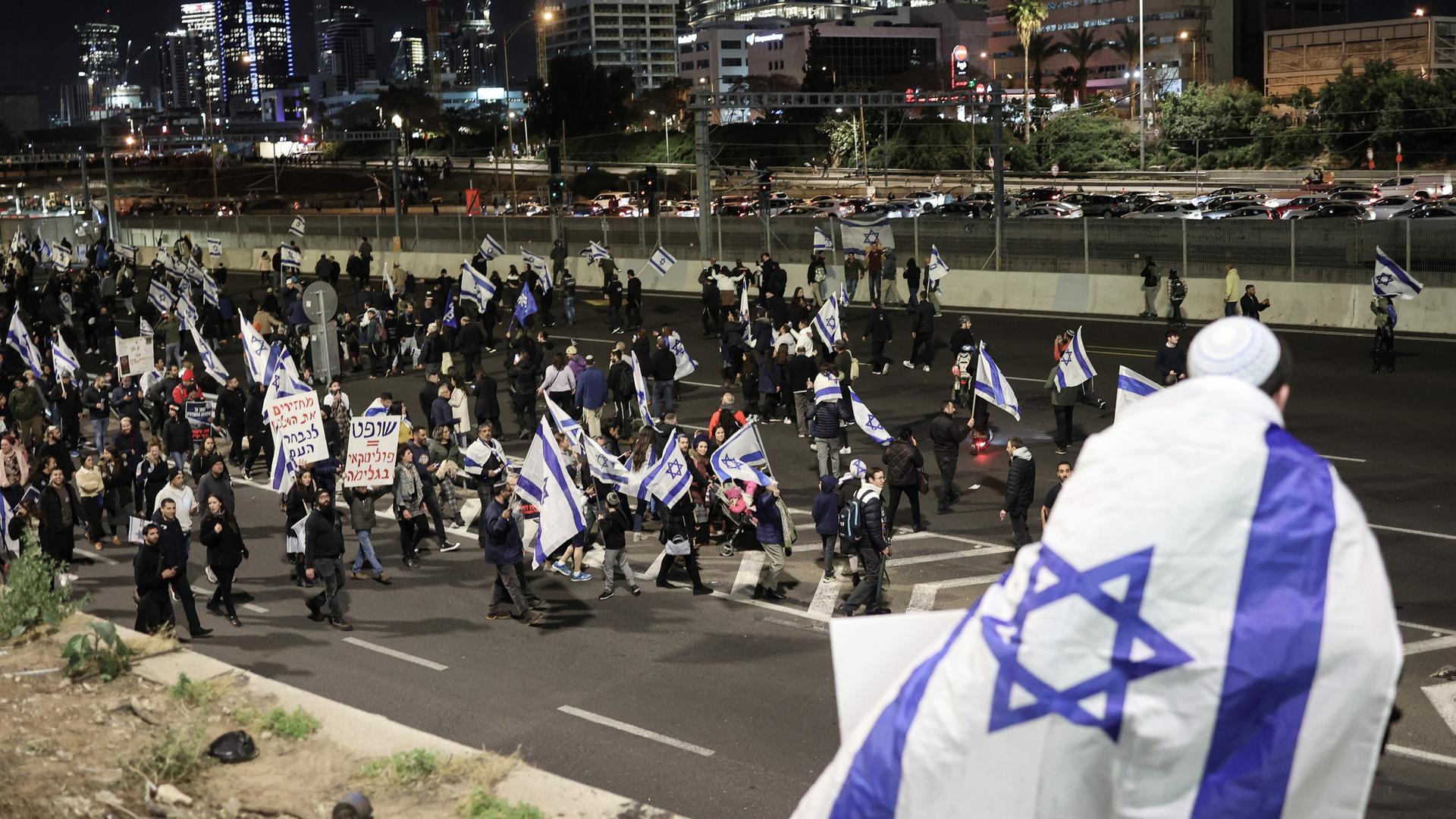 Nächtliche Szene in Tel Aviv, Israel: Menschen ziehen mit israelischen Fahnen und Transparenten durch die Stadt, im Vordergrund ist ein Mann mit Kippa, eingehüllt in eine große Fahne. Nächtliche Szene in Tel Aviv, Israel: Menschen ziehen mit israelischen Fahnen und Transparenten durch die Stadt, im Vordergrund ist ein Mann mit Kippa, eingehüllt in eine große Fahne.