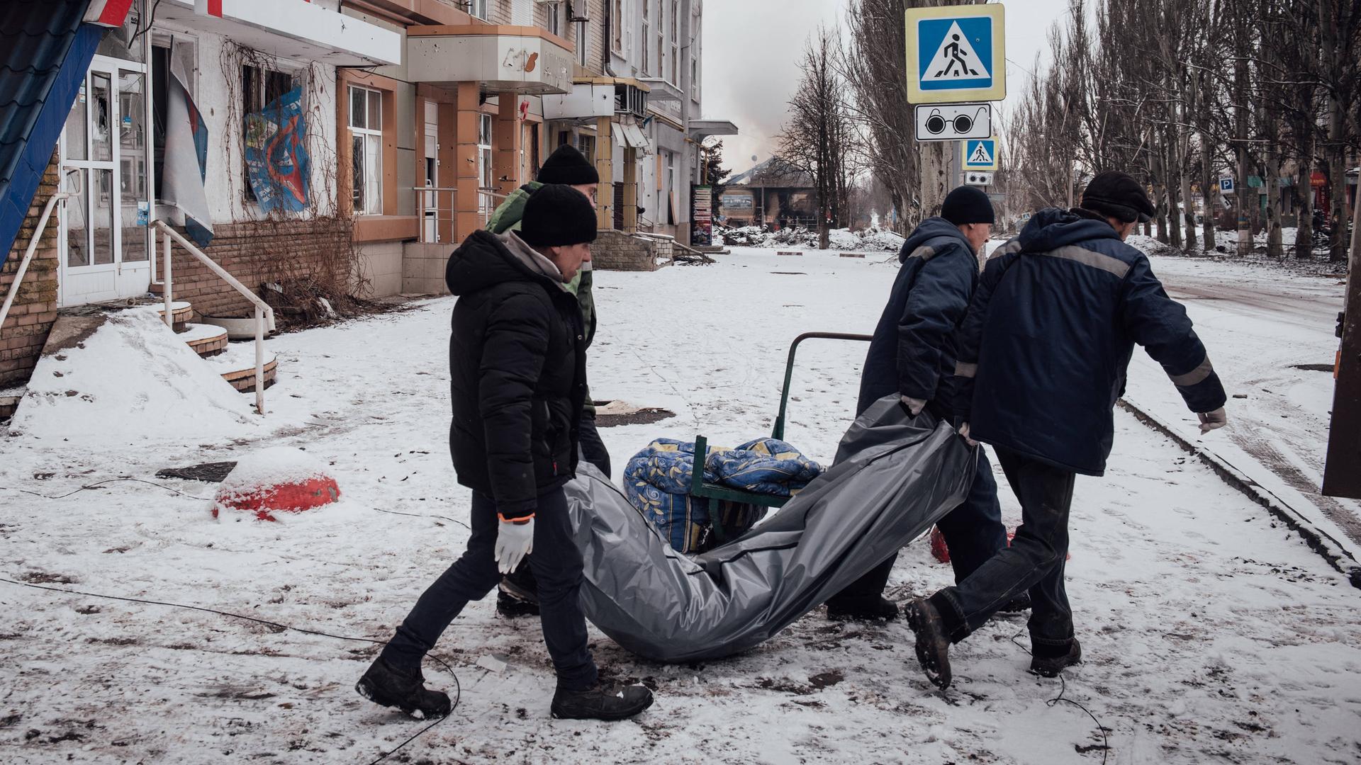 Vier Menschen tragen einen Leichensack durch eine verschneite Straße.