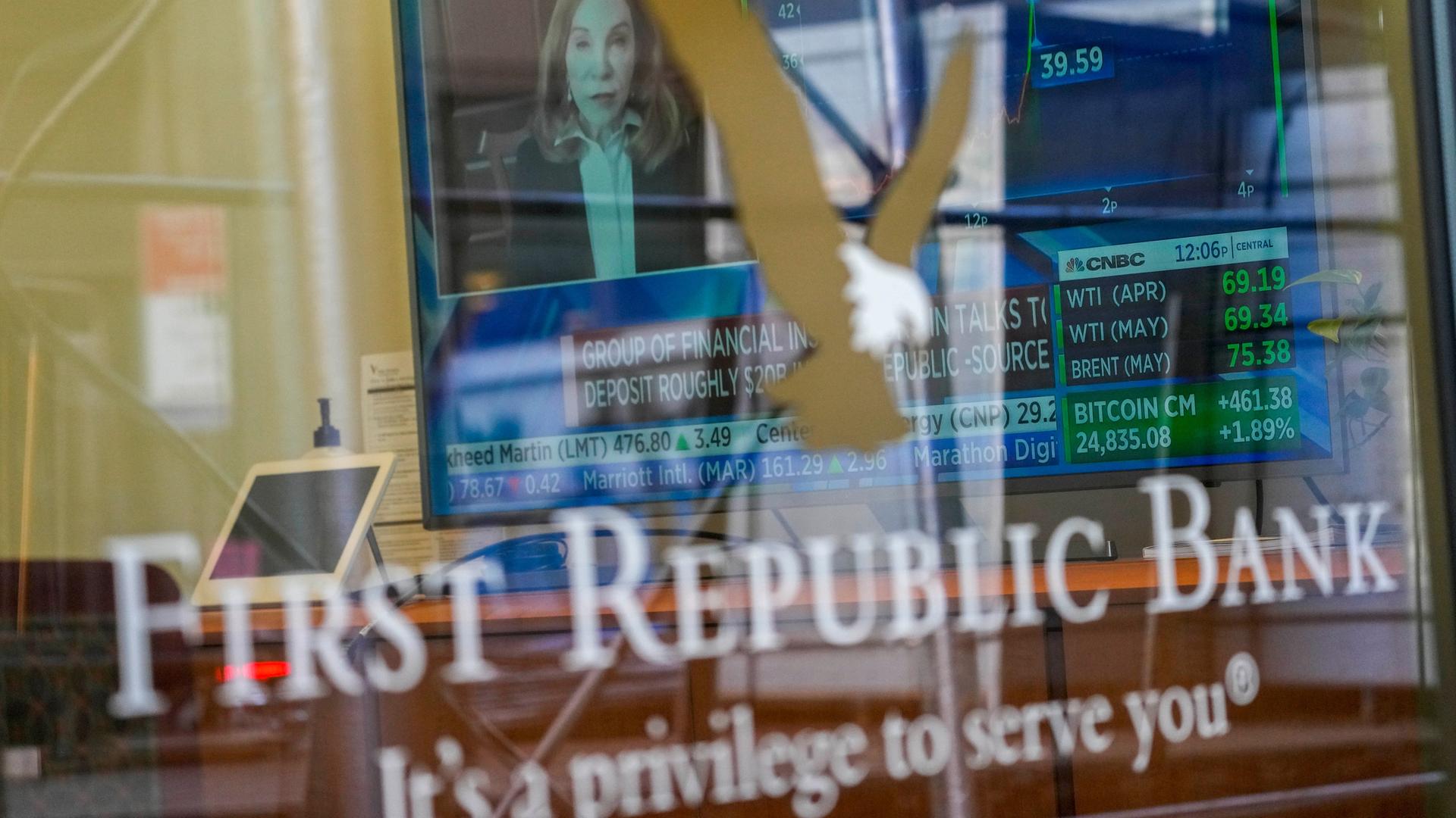 Blick auf die Scheibe, die eine Häuserwand spiegelt. Dahinter sieht man einen Bildschirm mit Finanzdaten einer Nachrichtensendung. Auf der Scheibe steht "First Republic bank - It´s a privilege to serve you"