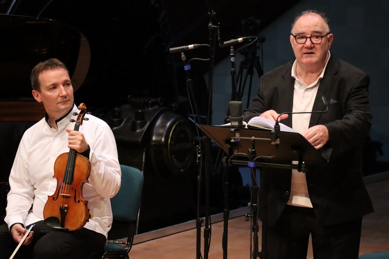 Ein Schauspieler steht in Konzertkleidung auf der Bühne und spricht in ein Mikrofon. Neben ihm sitzt ein Geiger mit seinem Instrument. Ein Schauspieler steht in Konzertkleidung auf der Bühne und spricht in ein Mikrofon. Neben ihm sitzt ein Geiger mit seinem Instrument.
