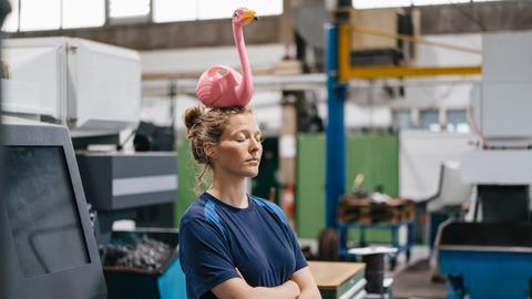 Eine Arbeiterin steht in einer Fertigungshalle mit verschränkten Armen und geschlossenen Augen. Auf ihrem Kopf balanciert ein pinker Plastikflamingo.