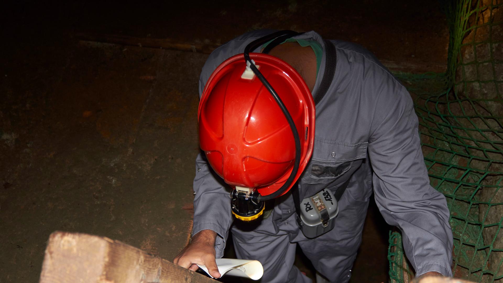 Ein Bergarbeiter mit rotem Helm und Stirnlampe steht auf einer Holztreppe und schaut hinunter in ein Bergwerk. 