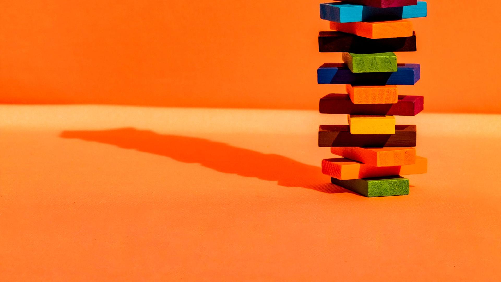 Ein Stapelturm aus bunten Bauklötzen vor orangefarbenem Hintergrund. Der Turm wirft einen kleinen Schatten auf der linken Seite. Ein Stapelturm aus bunten Bauklötzen vor orangefarbenem Hintergrund. Der Turm wirft einen kleinen Schatten auf der linken Seite.