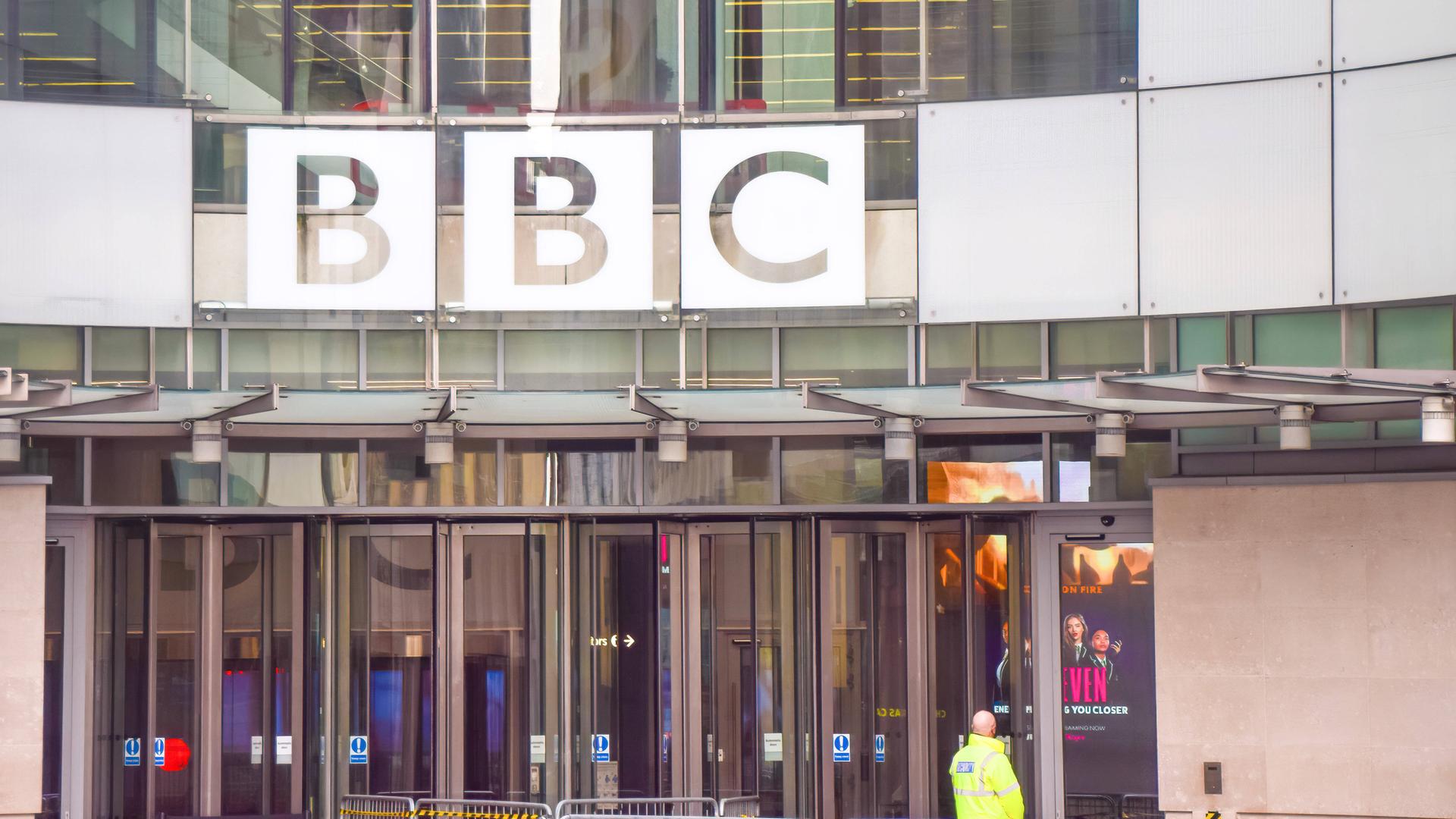 Eingangsbereich des BBC Broadcasting House im Zentrum von London. Über den gläsernen Eingangstüren ist der Schriftzug "BBC" zu sehen. Vor den Türen steht ein Sicherheitsmann in gelber Warnweste mit dem Rücken zur Kamera.