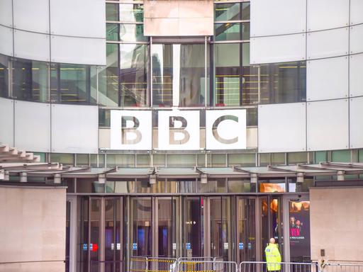 Eingangsbereich des BBC Broadcasting House im Zentrum von London. Über den gläsernen Eingangstüren ist der Schriftzug "BBC" zu sehen. Vor den Türen steht ein Sicherheitsmann in gelber Warnweste mit dem Rücken zur Kamera.
