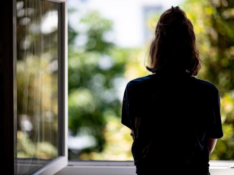Eine Frau steht in ihrer Wohnung an einem Fenster. 