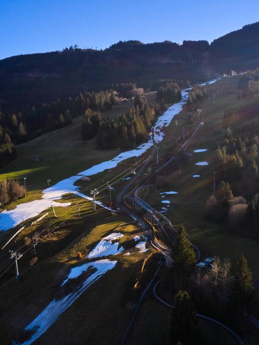 Blick von oben auf ein Alpenpanorama mit weißen Pisten-Streifen mit Kunstschnee umrahmt von grünen Wiesen wie in Nesselwang