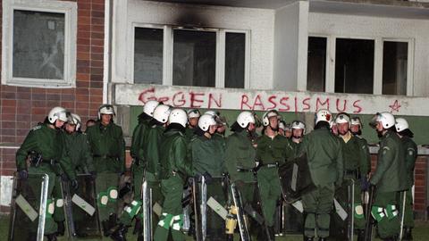 Polizisten stehen in Schutzuniform vor einem teils ausgebrannten DDR-Neubau. An den zersplitterten Fenstern sind schwarze Rauchspuren.