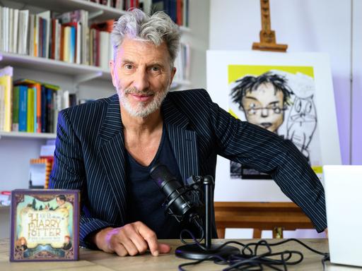 Der Schauspieler Rufus Beck sitzt an einem Tisch vor einer Bücherwand, links steht eine CD mit der Aufschrift "Harry Potter", rechts hinter Beck ist eine Zeichnung zu sehen, die Harry Potter darstellt, ein Kind mit einer Brille.