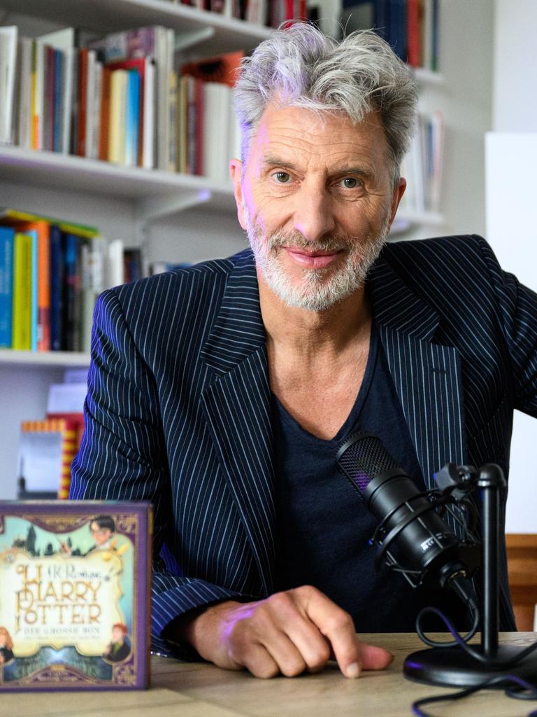 Der Schauspieler Rufus Beck sitzt an einem Tisch vor einer Bücherwand, links steht eine CD mit der Aufschrift "Harry Potter", rechts hinter Beck ist eine Zeichnung zu sehen, die Harry Potter darstellt, ein Kind mit einer Brille.
