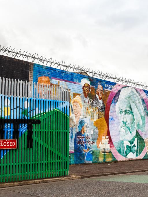 Wandbemalungen in der Northumberland Street, nahe der Falls Road in Belfast. Davor ein Tor mit der Aufschrift "Road closed", das eine sogenannte Peace Line (Friedenslinie) zur Trennung von Konfliktparteien darstellt. Abends wird das Tor geschlossen. 